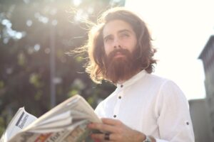 A younger man with brown hair and a beard is looking above the camera and holding a newspaper. He is outside and the sun is just rising behind a tree.